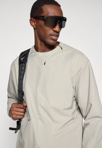 Veste gris clair, résistante à l'eau, avec un col haut et un design minimaliste ; le modèle porte de grandes lunettes de soleil noires et un sac à dos noir.
