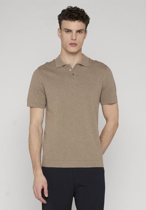 Junger Mann steht, trägt ein kurzärmeliges beigefarbenes Strick-Poloshirt und dunkle Hose vor einem schlichten hellen Hintergrund.