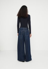 Mulher com cabelo encaracolado vestindo uma camisola preta de manga comprida e jeans largos de cintura alta azul escuro, em pé contra uma parede branca.