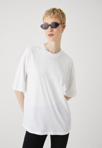 Tricou alb din bumbac cu mâneci scurte, decolteu rotund, croială lejeră, purtat cu pantaloni negri și ochelari de soare negri, subțiri și unghiulari.