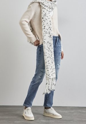 Personne portant un pull crème, une longue écharpe blanche mouchetée, un jean bleu usé et des baskets blanches, debout sur un sol gris.