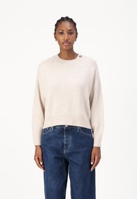 Beige avkortet genser med rund hals og ribbestrikket kant, med en knappdetalj på skulderen; kombinert med mørkeblå, løstsittende jeans.