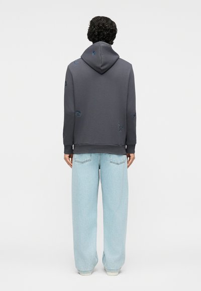 Άνδρας με πλάτη φοράει σκούρο γκρι φούτερ με μικρά κεντημένα μπαλώματα και ανοιχτό μπλε παντελόνι με φαρδιές μπατζάκια, πάνω σε απλό φόντο.