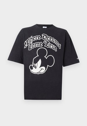 Mand iført sort oversized T-shirt med hvid Mickey Mouse-grafik og teksten "Where Dreams Come True" på fronten, der står foran en ensfarvet baggrund.