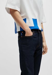 Camisa blanca de manga corta con un diseño geométrico azul en el dobladillo. Combinada con unos vaqueros de mezclilla azul oscuro, que presentan un corte y textura clásicos.