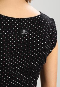 Rückansicht einer Person, die ein schwarzes Oberteil mit kleinen weißen und rosa Punkten trägt, sowie einem rechteckigen schwarzen Pflegeetikett nahe dem Ausschnitt.