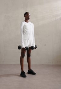 White long-sleeve athletic top and shorts, holding black dumbbells, standing on gray flooring against a light concrete wall.