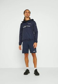 Tumedane tuulejope, millel on silmapaistev logo, kapuuts ja elastne alumine äär. Koos sellega on sobivad tumesinised lühikesed püksid ja mustad spordijalatsid.