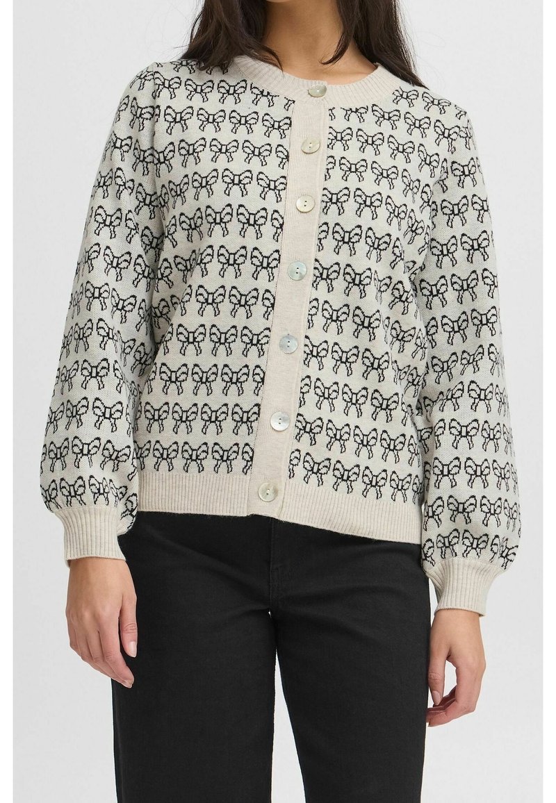 Femme portant un cardigan crème boutonné avec des motifs de nœuds noirs et des manches bouffantes, assorti à un pantalon noir sur fond blanc.