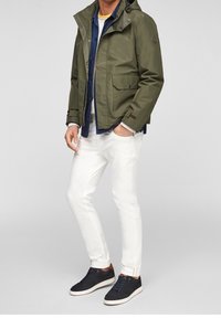 Uomo che indossa una giacca a hood verde oliva, una camicia blu navy, pantaloni bianchi e sneakers nere, in piedi con una mano in tasca.