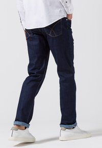 Jeans de mezclilla azul oscuro con corte recto, con costuras en color beige, un clásico diseño de cinco bolsillos y puños enrollados. Combinados con zapatillas blancas.