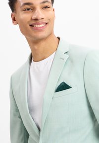 Heller Mintblazer aus glattem Stoff, mit einem Reverskragen, einer einzelnen Tasche mit einem grünen Einstecktuch, getragen über einem weißen Hemd.