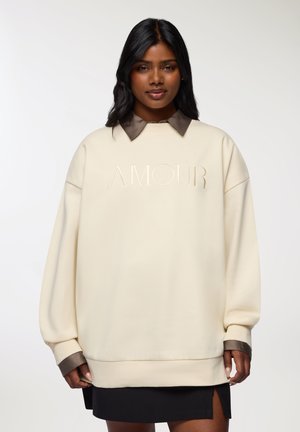 Felpa oversize color crema con colletto marrone, caratterizzata dalla scritta "AMOUR" in rilievo sul davanti e polsini e orlo a costine.