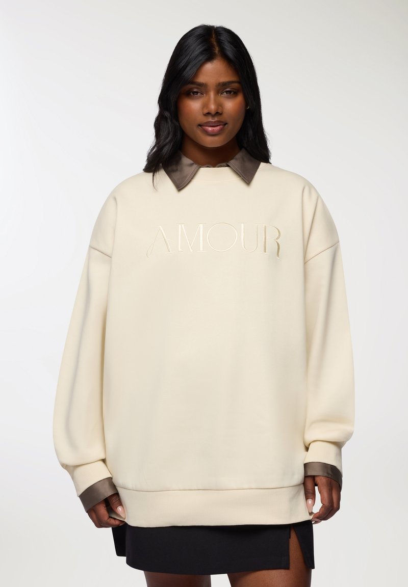 Felpa oversize color crema con colletto marrone, caratterizzata dalla scritta "AMOUR" in rilievo sul davanti e polsini e orlo a costine.