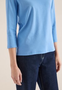 Personne portant une chemise à manches longues bleu clair et un jean bleu foncé, debout avec les bras détendus le long du corps devant un fond neutre.