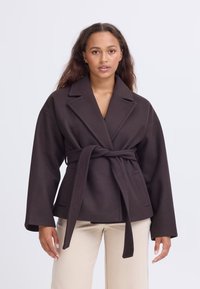 Donna con lunghi capelli castani che indossa un cappotto scuro belato con cintura e pantaloni beige chiaro su uno sfondo chiaro e semplice.