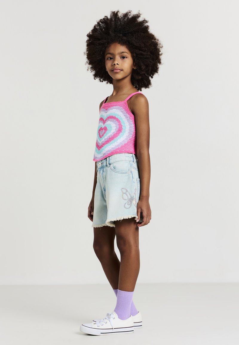 Häkel-Top in pinkem und blauem Herzmuster, kombiniert mit hellblauen Denim-Shorts mit fransigem Saum und Schmetterlingsstickerei. Weiße Sneaker, lila Socken.