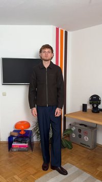 Man som står inomhus iklädd en mörk jacka och marinblå byxor, med en TV, en orange lampa, böcker och en hylla med ett headset och en resväska i närheten.