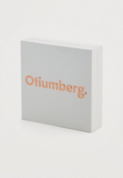 Τετράγωνο γκρι κουτί με λεία υφή και λογότυπο "Otiumberg." σε ροζ χρυσό μπροστά. Περιέχει καθαρό, μοντέρνο σχεδιασμό.