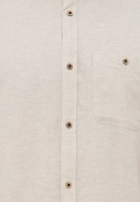 Chemise beige clair avec des boutons en bois sur le devant et une poche poitrine avec un bouton assorti.
