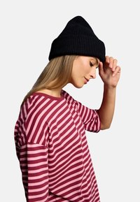 Black knit beanie with a ribbed texture, worn by a person in a burgundy and pink striped shirt, shown in profile against a neutral background.