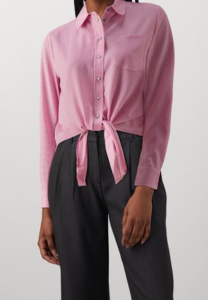 Kobieta ubrana w różową koszulę w paski zapinaną na guziki, wiązaną w talii, z długimi rękawami oraz ciemne spodnie z wysokim stanem.