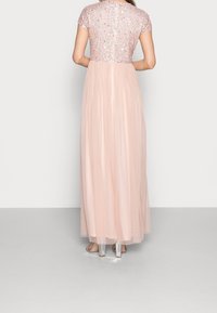 Vestido maxi con un cuerpo de manga corta y lentejuelas en rosa claro, falda de tul fluida y tacones transparentes. La vista trasera muestra el detalle de la cremallera.