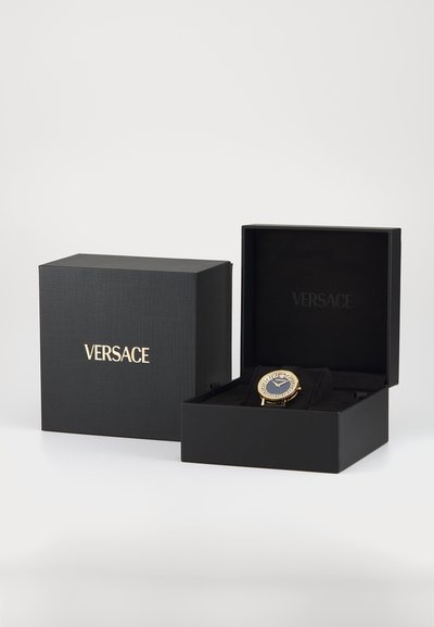 Črna škatla z zlatim logotipom "Versace", ki razkriva zlato uro z okrasnim modrim številčnicom in zapletenimi podrobnostmi na pasu, postavljena na črno oblazinjenje.