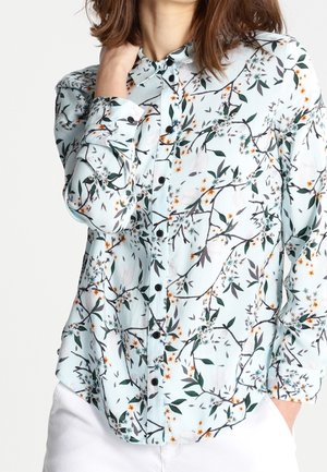 Vrouw draagt een lichtblauwe blouse met knopen en een bloem- en takkenpatroon, gecombineerd met een witte broek, hand rust dichtbij de hals.