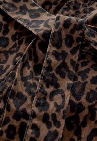 Tissu imprimé léopard en marron et noir avec une texture douce. Présente des coutures visibles et un détail en ruban, mettant en valeur le design.