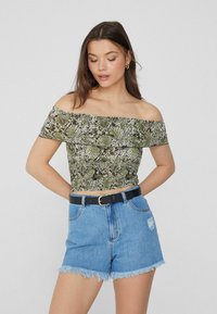 Crop top off-shoulder in stampa serpente verde e nera, abbinato a pantaloni shorts in denim a vita alta con orlo sfrangiato e una cintura nera.