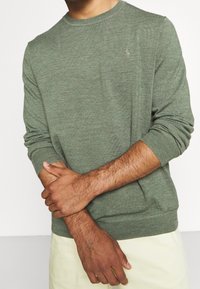 Man som bär en långärmad grön tröja med en liten broderad logotyp, med armarna korsade vid handlederna, och ljusa byxor.