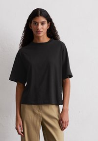 Maglietta nera a maniche corte in cotone, con una vestibilità comoda e colletto rotondo, abbinata a pantaloni beige a vita alta.
