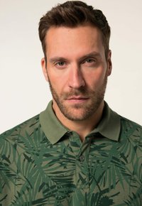 Grünes Poloshirt mit Blättermuster, aus weichem Baumwollmaterial. Es verfügt über einen Kragen und Knopfdetails am Hals.