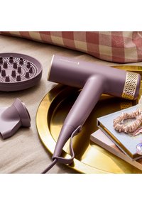 Lilla hårføner med gull detaljer, glatt matt overflate, tilkoblet ledning, diffuser og konsentratordyse på et gullfat blant tilbehør.