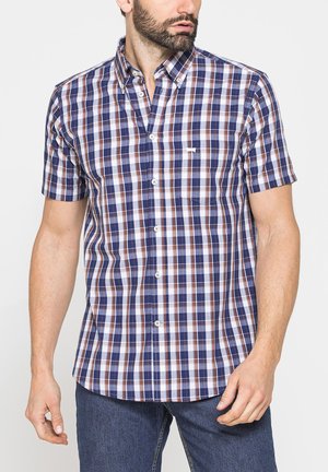 Uomo che indossa una camicia a maniche corte a quadri blu, bianca e marrone con taschino sul petto e jeans blu, rivolto leggermente verso sinistra.