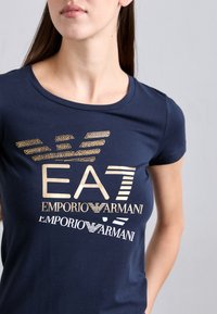 Tamsiai mėlyna trumpomis rankovėmis marškinėliai su metaliniu auksiniu tekstu ir logotipų dizainu, apvaliu iškirpimu ir minkšta medvilnės tekstūra.