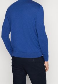 Maglione blu a girocollo con maniche lunghe, polsini e orlo a costine. Texture liscia, design aderente. Indossato sopra pantaloni scuri.