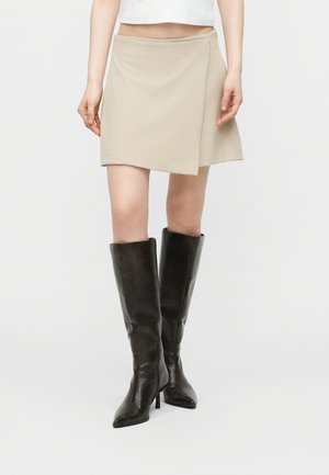 Femme portant une mini jupe portefeuille beige associée à des bottes noires montantes à talons hauts et bouts pointus, debout devant un fond blanc.