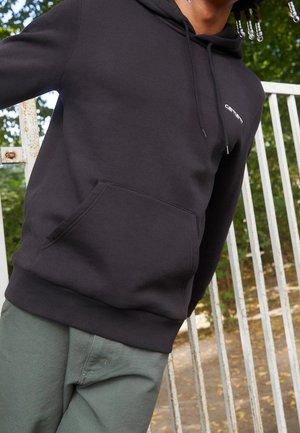 Person trägt einen schwarzen Hoodie mit "Carhartt"-Logo, steht draußen neben einem weißen Metallzaun mit grünen Pflanzen im Hintergrund.