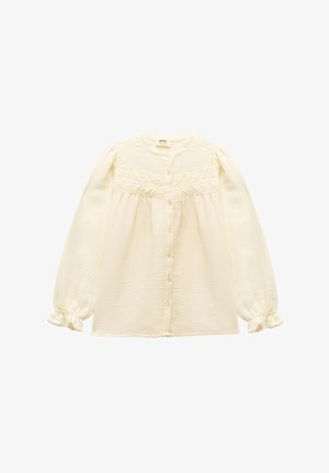 Valkoinen napillinen paita, jossa on pitkät, puffatut hihat, koottuja yksityiskohtia ja röyhelöalaset. Valmistettu kevyestä, rakenteellisesta kankaasta.