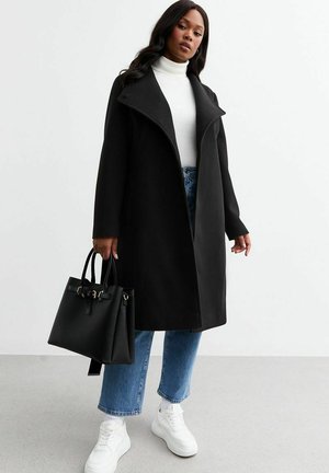 Zwarte wollen jas met hoge kraag, gecombineerd met een witte coltrui, blauwe wijde jeans en witte sneakers. Houd een zwarte handtas vast.