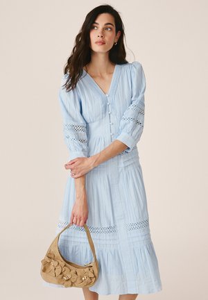 Kobieta ubrana w jasnoniebieską sukienkę midi z bufkami i koronkowymi detalami, trzyma beżową kwiatową torebkę z koralikami.