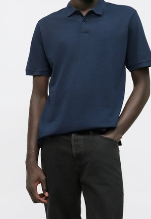 Homme portant un polo bleu marine à manches courtes et un jean noir, une main dans la poche et l'autre reposant à ses côtés.
