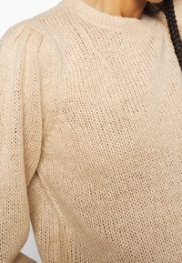 Beige neulottu neulepaita, jossa pyöreä pääntie, teksturoitu avoin kudonta ja pullistetut hihat, pehmeästä ja kevyestä materiaalista.