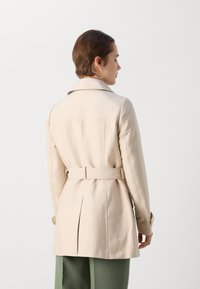 Beige trenchcoat met een met riem verstelbare taille, achterventilatie en brede kraag. Heeft een gladde textuur en lange mouwen.
