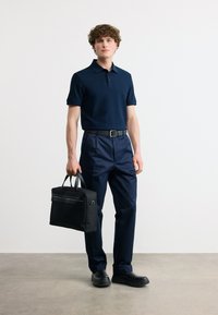 Άνδρας που φοράει μπλε ναυτικό πόλο πουκάμισο και παντελόνια, μαύρη ζώνη και παπούτσια, κρατώντας μια μαύρη τσάντα εργασίας, στέκεται σε απλό φόντο.