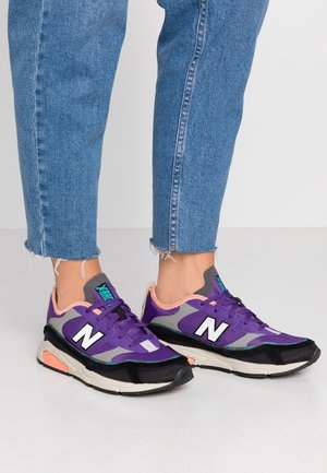 Fioletowe sneakersy New Balance z szarymi i czarnymi akcentami, posiadające teksturowaną cholewkę, sznurowadła oraz amortyzowaną podeszwę z pomarańczowymi detalami.