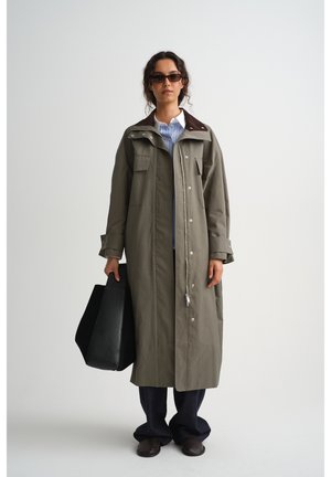 Frau mit langem olivgrünem Mantel, gestreiftem Hemd, dunkler Hose, Sonnenbrille, die eine große schwarze Tragetasche hält und vor einem schlichten Hintergrund steht.