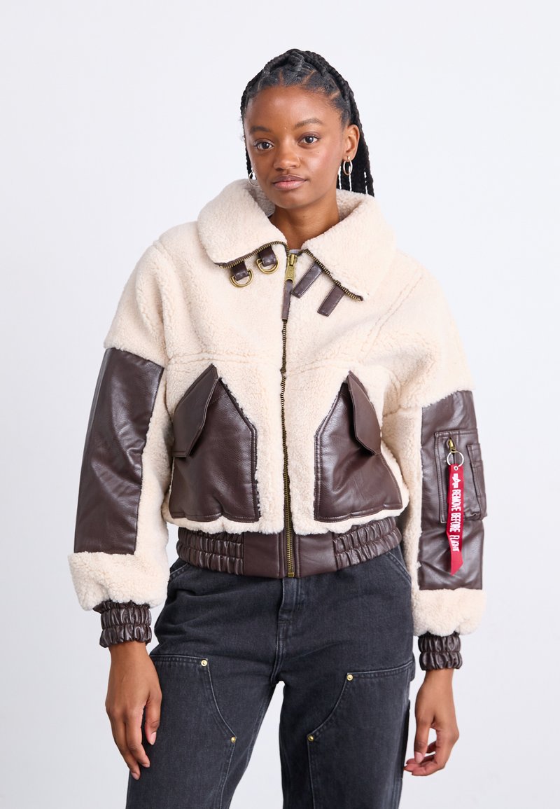 Giacca in pelle marrone e finta shearling crema con colletto alto, due grandi tasche frontali, polsini elastici e chiusura a zip. Dettaglio con etichetta rossa.